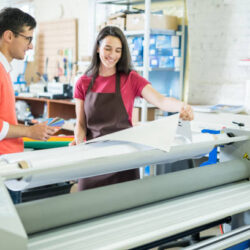 Cheerful excited beautiful lady in apron showing printed banner to manager who picking out color on swatch for printing